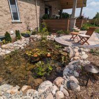 Small pond, surrounded by rocks, mulch, outdoor lights, and shrubs. There is a flagstone patio with a wooden chair and table nearby.