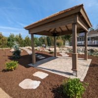 Pergola with custom stone patio underneath, surrounded by mulch, stone walkway, and shrubs.